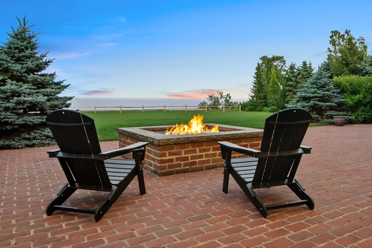 Adirondack chairs around a glowing brick fire pit with a lake view at sunset